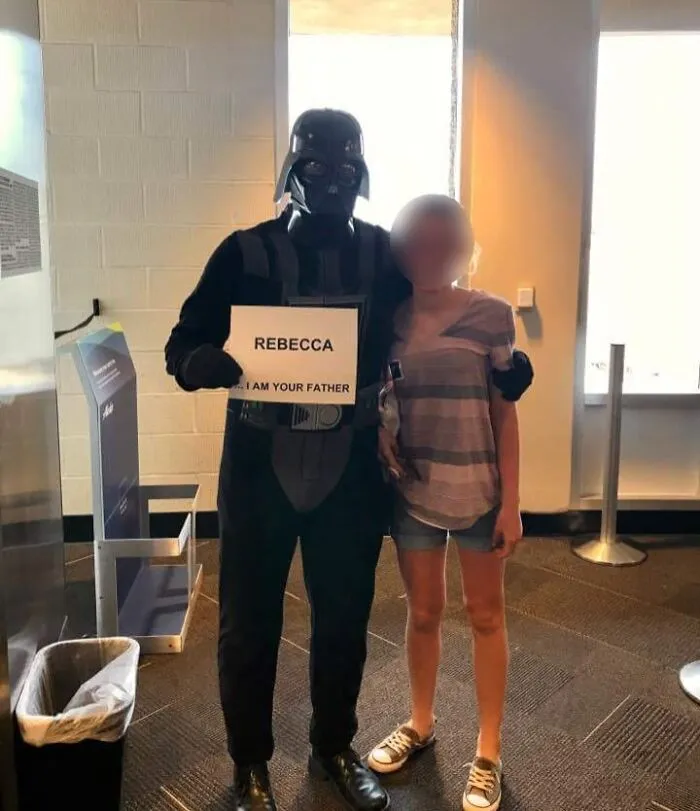 Osoba v kostýme s nápisom stojí vedľa inej osoby vo vnútri budovy.