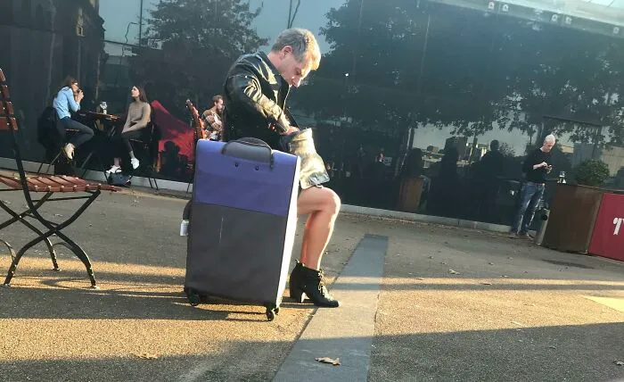 Osoba sedí na kufri na vonkajšej terase, zatiaľ čo si obúva topánku. V pozadí ľudia pri stoloch.