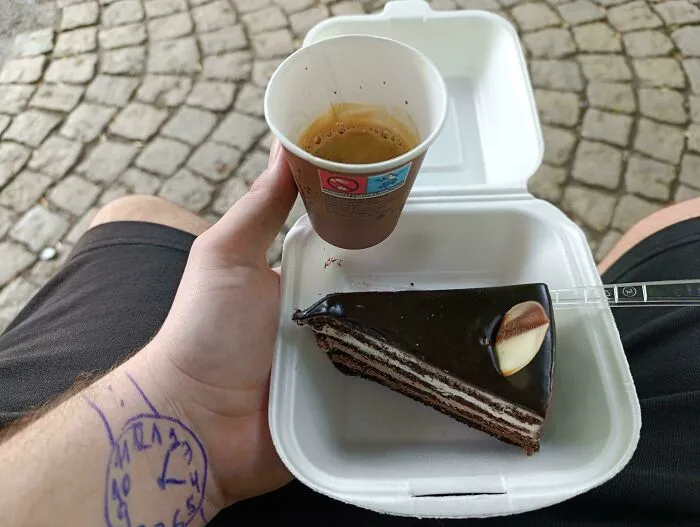 Osoba drží kávu a čokoládový zákusok na terase s dlažobnými kockami; ruka s nakreslenými hodinkami.