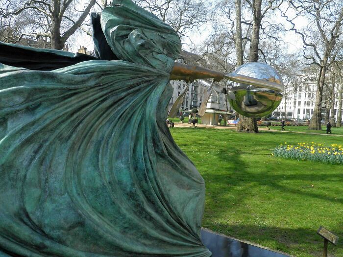 Socha ženy s vejárom a guľou na parku, umelecké dielo pod holým nebom medzi stromami.