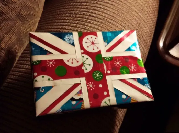 Darček zabalený v pestrofarebnom vianočnom papieri s britským vzorom, položený na hnedej pohovke.