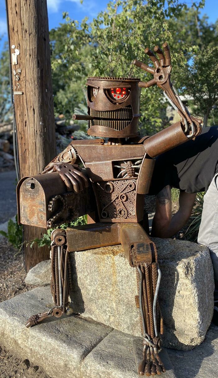 Kovová robotická socha so schránkou sedí na kameni, jeden zvedený pazúr pozdravuje.