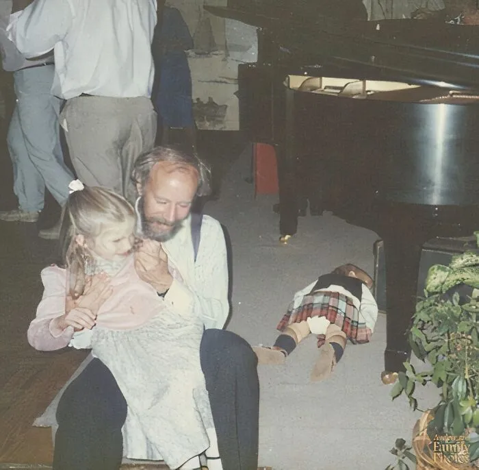 Otec objíma dcéru pri klavíri, zatiaľ čo ďalšie dieťa leží pri klavíri na zemi. Rodinná chvíľa.