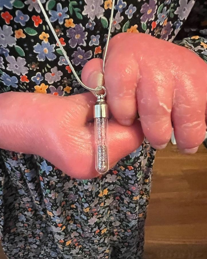 Náhrdelník s fľaštičkou obsahujúcou trblietky držanou rukou na kvetinových šatách.