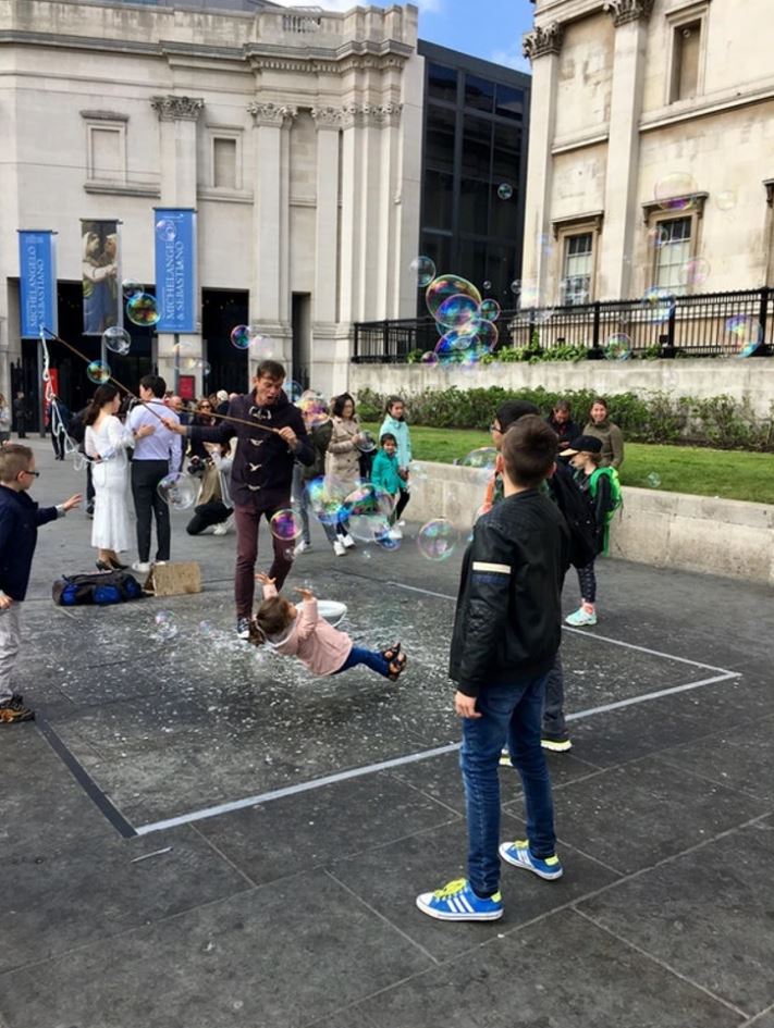 Deti sa hrajú s bublinami na rušnom námestí pred historickou budovou za slnečného dňa.