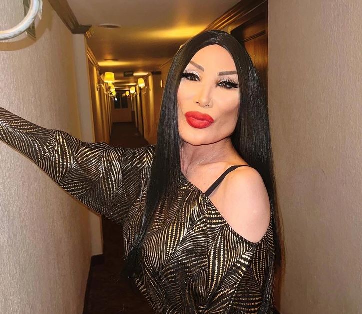 Osoba v šate s metalickým vzorom stojí v osvetlenej hotelovej chodbe a pózuje s úsmevom.