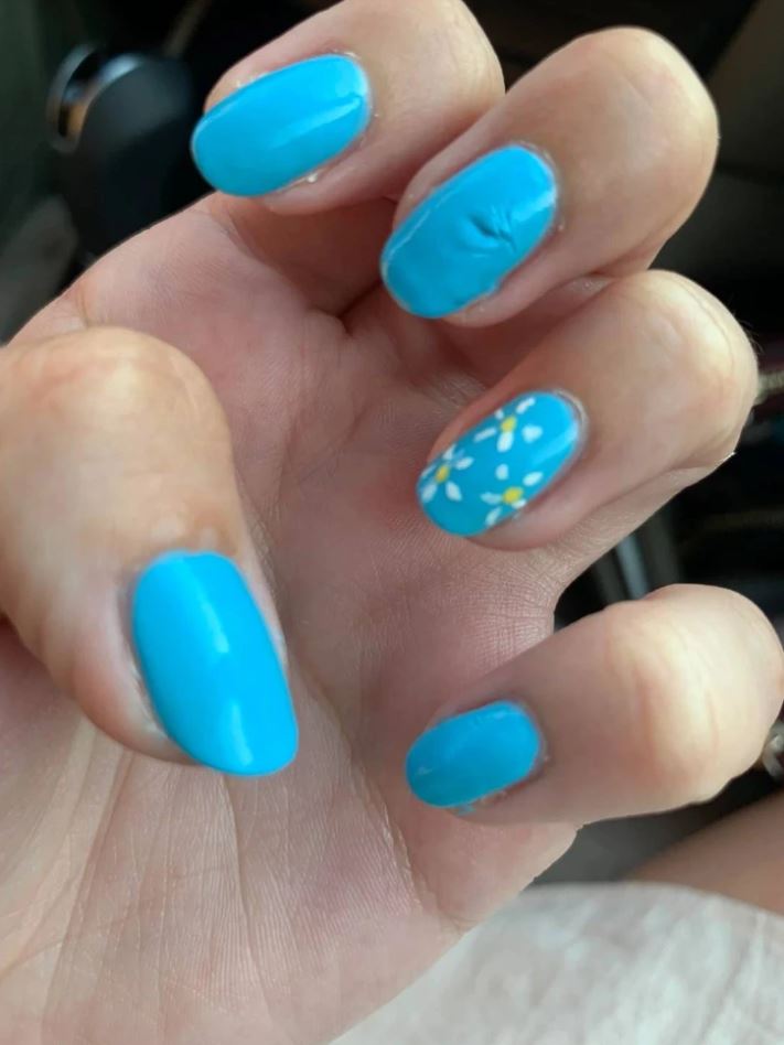 Ruka s modrým lakom na nechtoch a kvetinovým vzorom na jednom nechte.