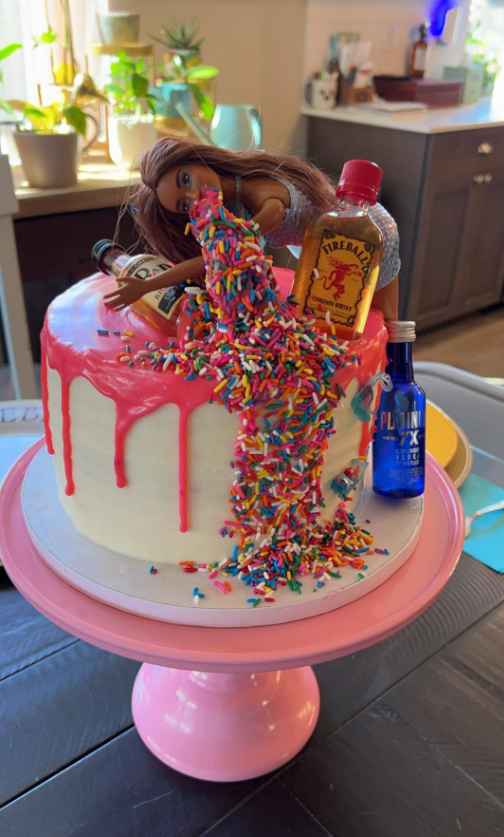 Ozdobená torta s bábikou, farebnými posýpkami a miniatúrnymi fľašami na ružovom podnose v kuchyni.