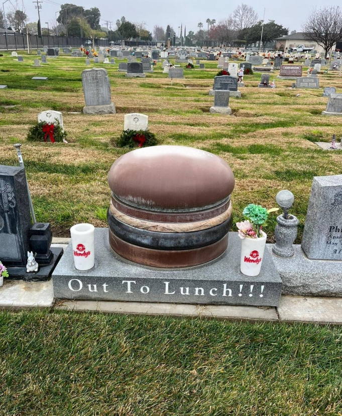 Náhrobný kameň v tvare hamburgeru s nápisom „Out To Lunch“ na cintoríne, vedľa pohárov Wendy’s.