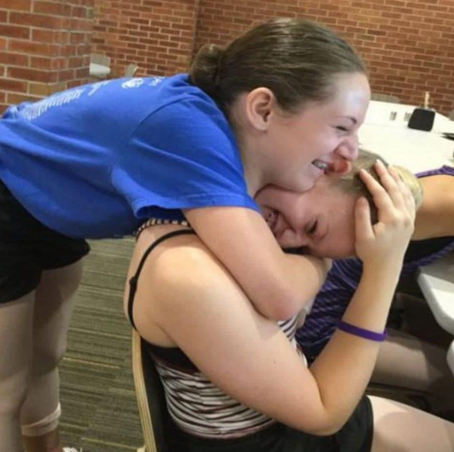 Dve dievčatá sa smejú a objímajú v miestnosti s tehlovou stenou, vyžarujú radosť a priateľstvo.