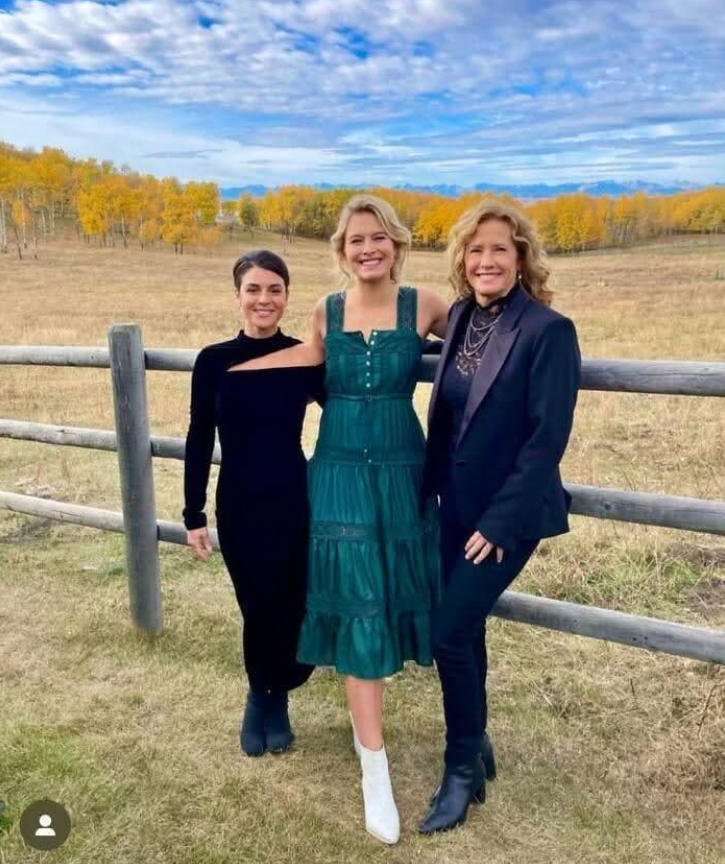 Neznáme osoby pózujú v jesennej krajine pred plotom so žltými stromami na pozadí.