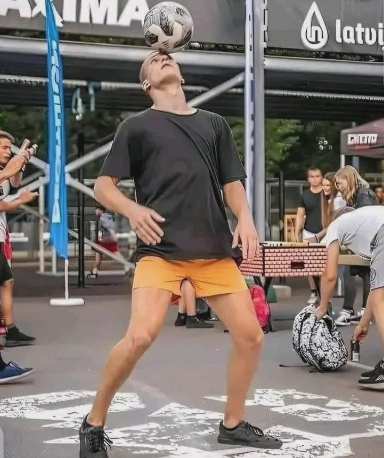 Muž v čiernom tričku a oranžových šortkách balansuje futbalovú loptu na čele na verejnej akcii.