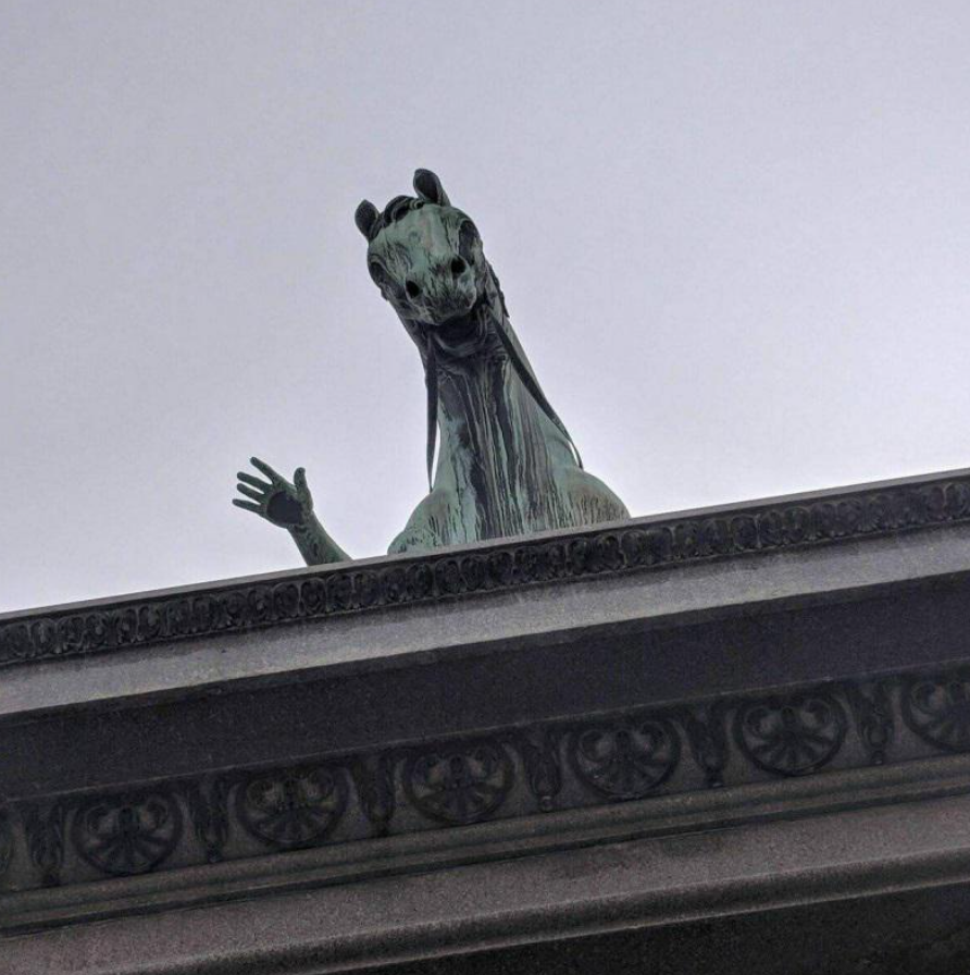 Socha koňa s komickou tvárou a vztýčenou rukou, pohľad zospodu na ozdobnom podstavci proti bledému nebu.