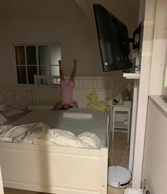 Dieťa stojí na hlave na posteli v izbe, vedľa je žltá hračka a na stene veľká zapnutá televízia.