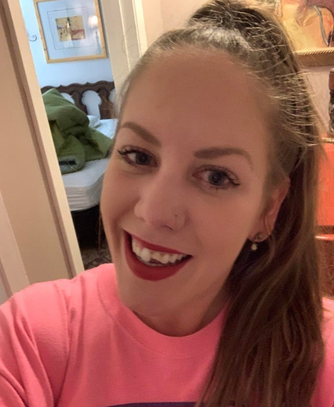 Usmievajúca sa žena v ružovom tričku s dlhou hnedou vlasmi vo vnútri, v pozadí vidno posteľ a obrazy.