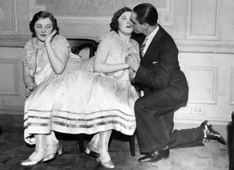 Dve ženy v retro šatách, jedna vyzerá zamyslene, druhá dostáva pusu od muža v obleku. Hallova scéna lásky.