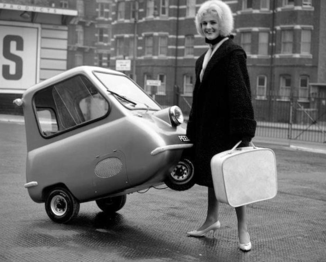 Žena stojí vedľa malého auta Peel P50, držiac kufor na ulici, 60. roky.