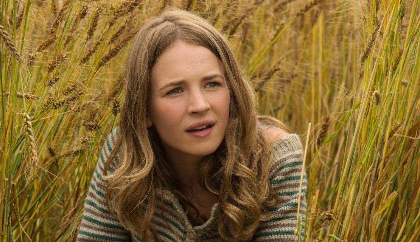 Žena v pruhovom svetri sedí v poli so zlatými klasmi, zamyslene sa pozerá do diaľky.