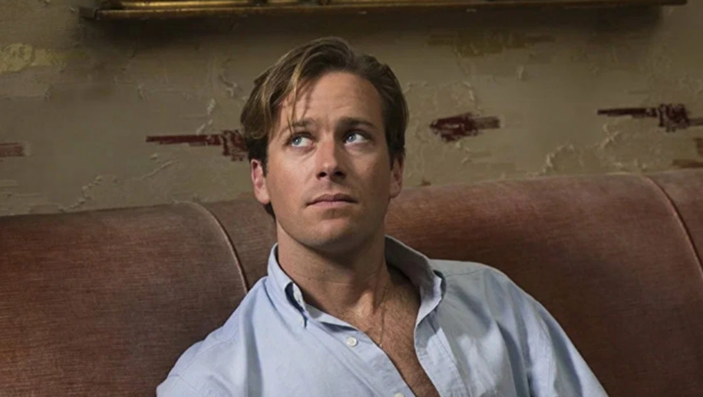 Osoba s modrou košeľou sedí na hnedej pohovke a pozerá sa do strany s vážnym výrazom.