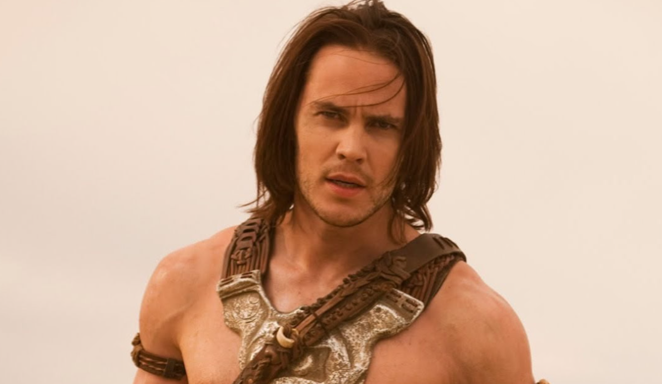 Muž v bojovom obleku s dlhými vlasmi na otvorenom priestranstve, pohľad smerom k fotoaparátu.