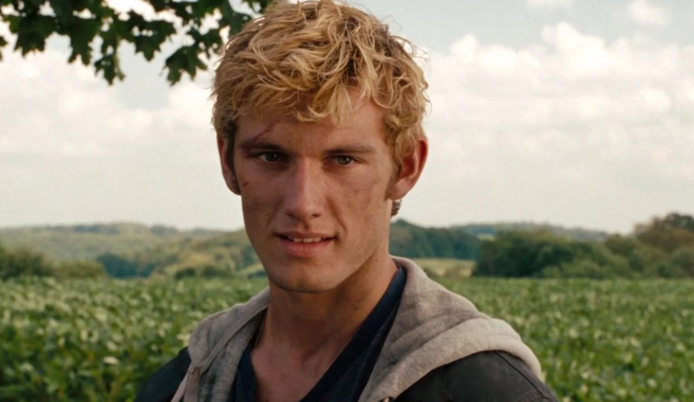 Osoba s blond vlasmi v prírode, pozerá jemne do strany. Pozadie tvoria zelené polia a modrá obloha.