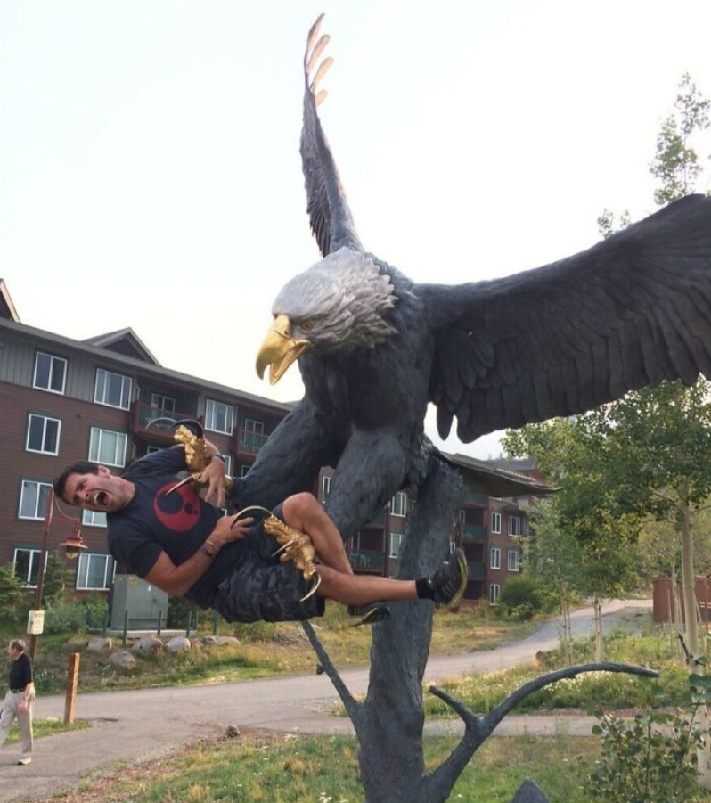 Ľudia pózujúci na soše obrovského orla pred budovami v pozadí, vtipná interakcia s umením.