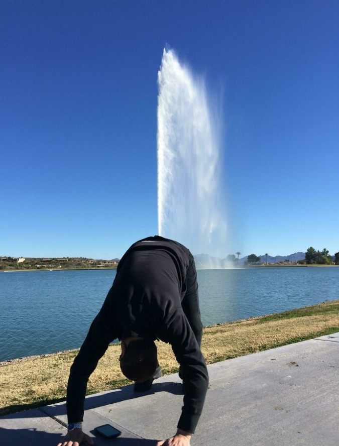 Osoba pred vodotryskom na brehu jazera v predklone, slnečný deň, modrá obloha.