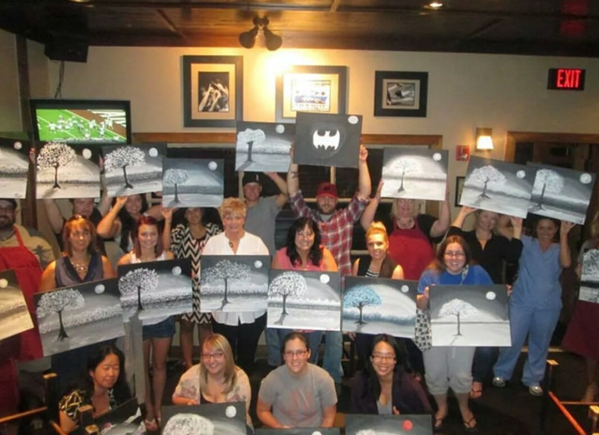 Skupina ľudí drží obrazy s motívom stromov na výtvarnom workshope v miestnosti s obrazmi a televíziou na stene.