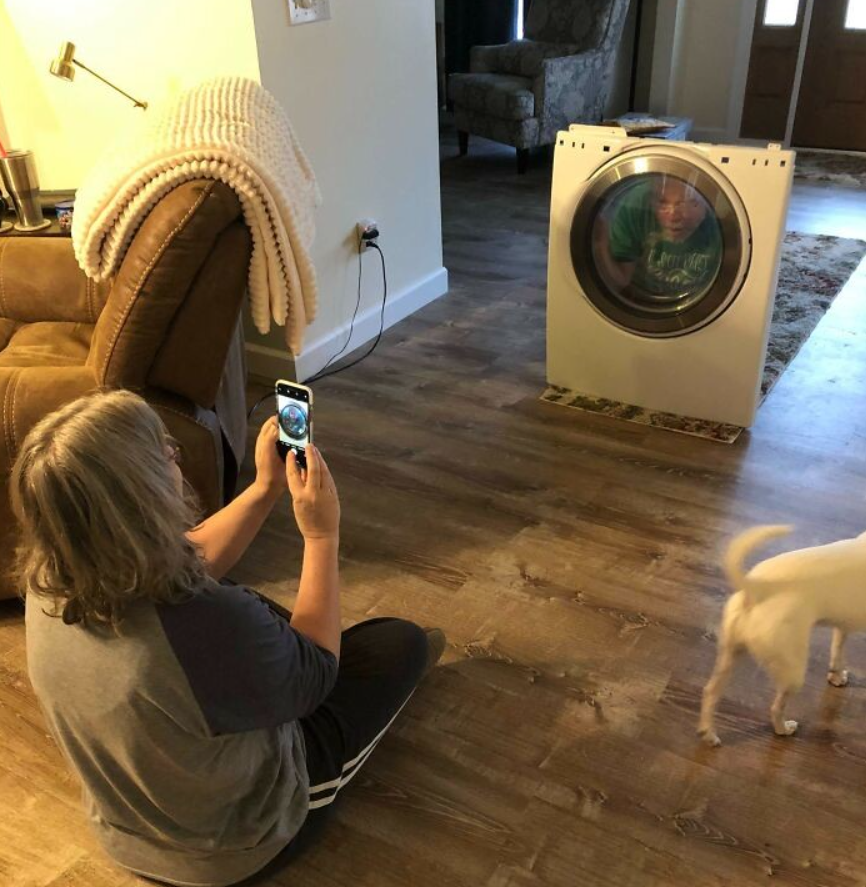 Osoba fotografuje dieťa sediace vo vybratom bubne práčky, zatiaľ čo prechádza malé psíča.