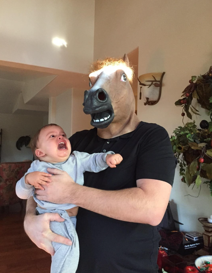 Muž s maskou koňa drží plačúce dieťa v obývačke, humoristická situácia.