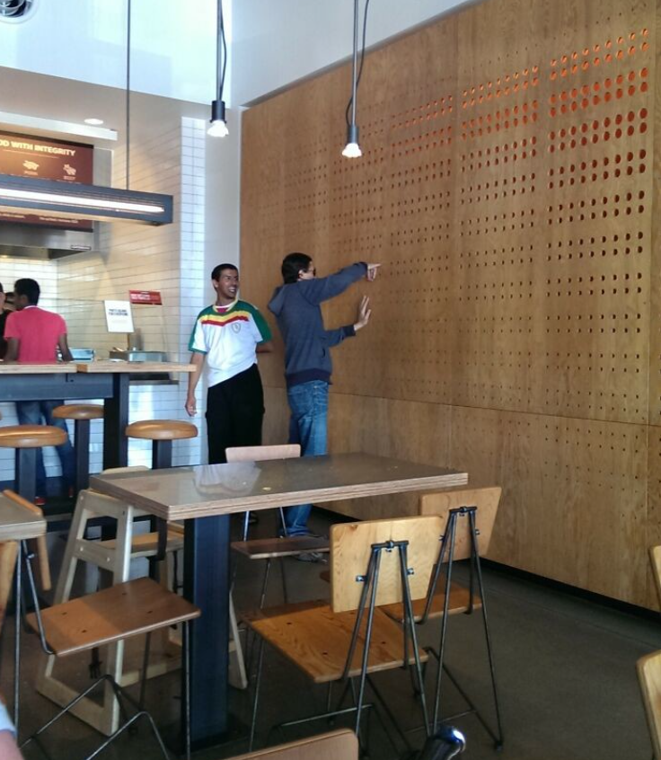 Dvaja muži v neformálnom oblečení gestikulujú pred drevenou stenou v modernej reštaurácii.