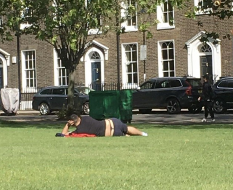 Osoba leží na tráve v mestskej záhrade, relaxuje pod stromom s radom domov v pozadí.