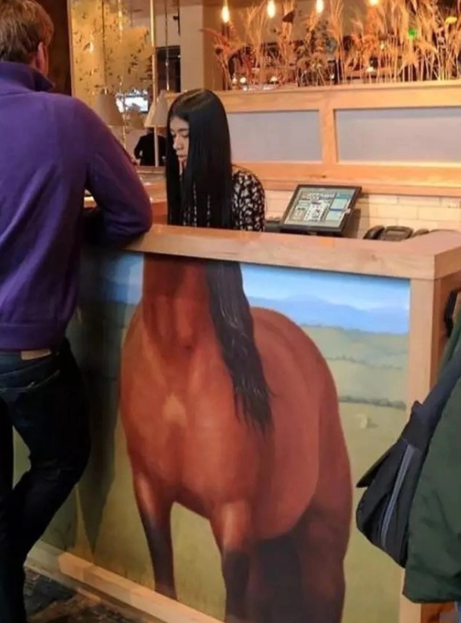 Mladá žena za pultom v kaviarni, umiestnená za maľbou koňa, vytvára ilúziu spojenia s jeho telom.