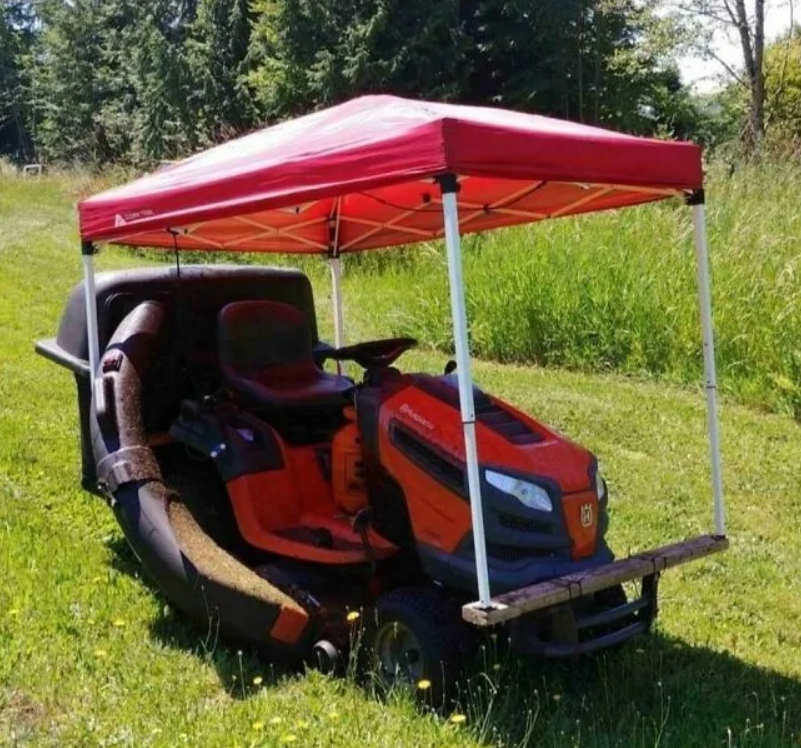 Kosačka pod červeným prístreškom na zelenej lúke, pripravená na kosenie trávy v slnečný deň.