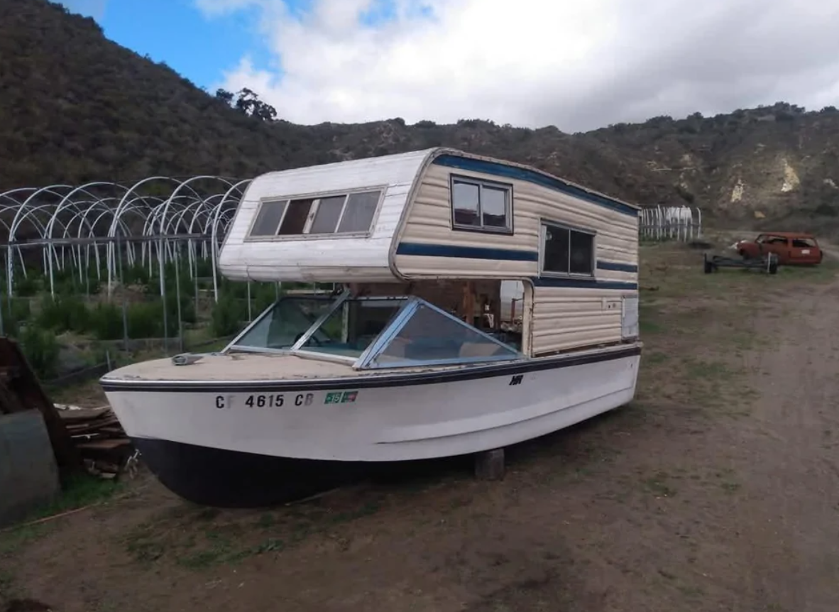 Loď s obytnou nadstavbou na súši, pozadie tvorí krajina s kopcami a fóliovníkami.
