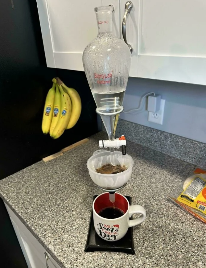 Chemické zariadenie používajúce lievik na prípravu kávy v kuchyni, banány a pohár na stole.