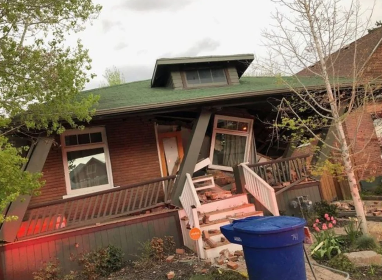 Poškodený dom s nakloneným verandovým stĺpom, tehlové múry v páde, modrý kontajner na odpad v popredí.