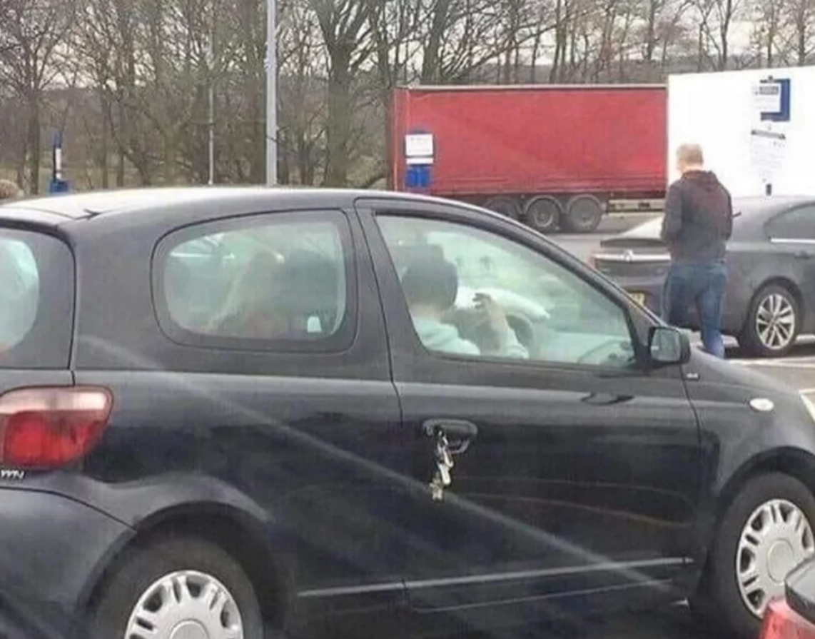 Čierne auto s kľúčmi zabudnutými v zámke; zamyslený chodec vzadu na parkovisku.