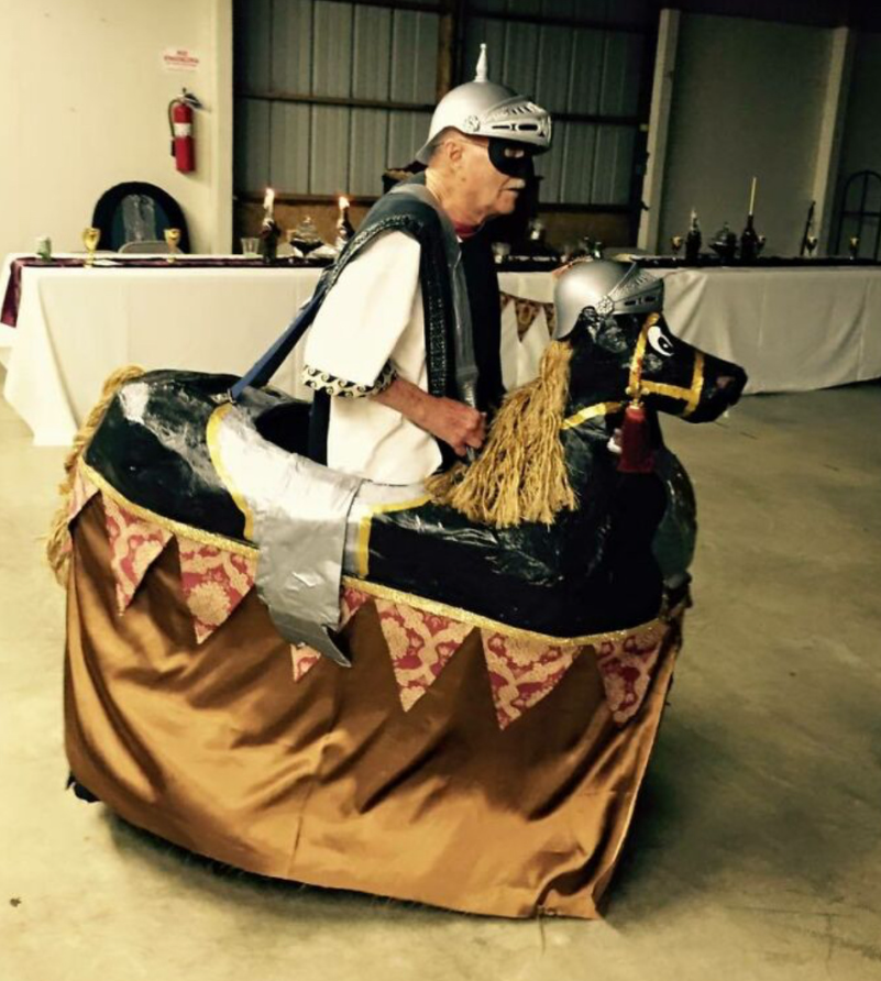 Muž v kostýme rytiera s kartónovým koňom na gurmánskom podujatí s dekoratívne prestretým stolom v pozadí.