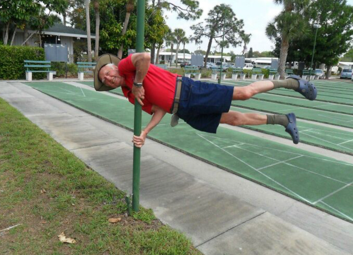 Muž v červenom tričku visí horizontálne na stĺpe v parku, oblečený v klobúku, ponožkách a modrých kroksách.