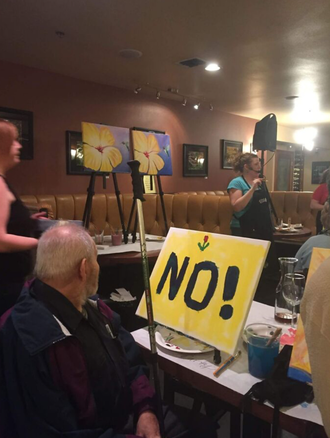 Starší pán na maľovaní: Na plátne žltý nápis NO! s kvetom v spoločenskej miestnosti.