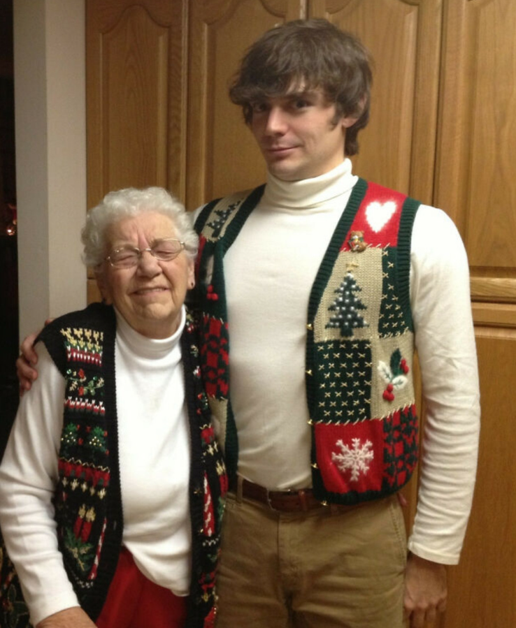 Dvojica ľudí v zladených vianočných vestách stojí pred drevenou skriňou, staršia žena s úsmevom zatvára oči.