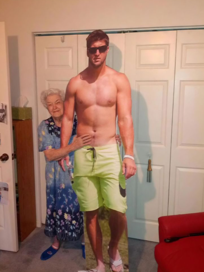 Staršia pani objíma kartónovú postavu muža v plavkách a slnečných okuliaroch pred bielymi dverami.