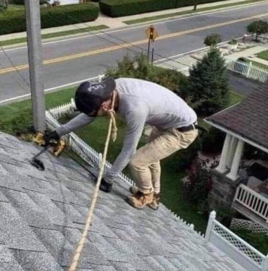 Muž pracuje na streche, drží sa lanom, používa vŕtačku na upevnenie škridiel.