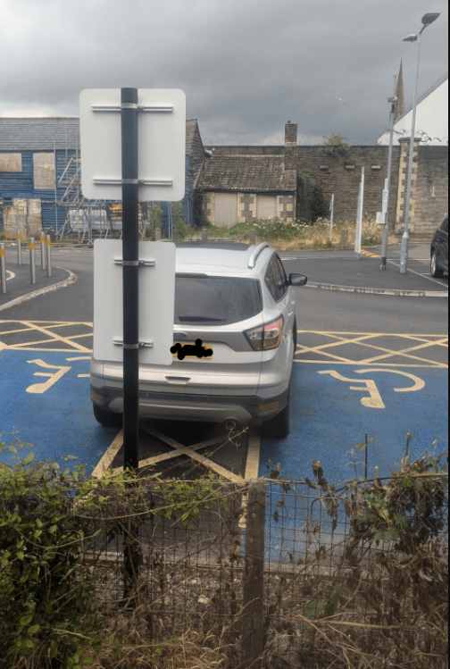 Auto zaparkované cez dve miesta pre telesne postihnutých na modrom povrchu, vzadu zabránené dopravným značkou.