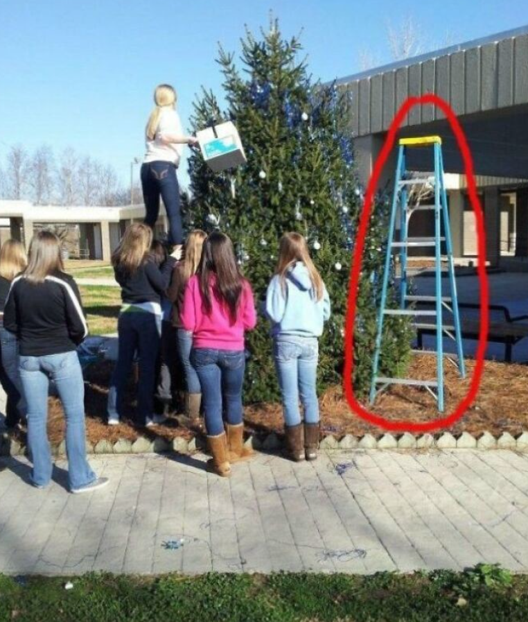 Skupina dievčat zdobí vianočný stromček kaskádovo použitou stoličkou, kým rebrík zostáva nevyužitý vedľa nich.