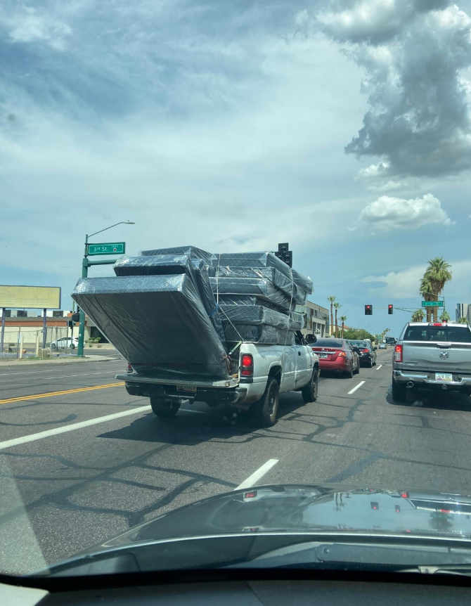 Nákladné auto na ceste nesúce veľké množstvo matracov, zabezpečené plastom pod modrou oblohou.