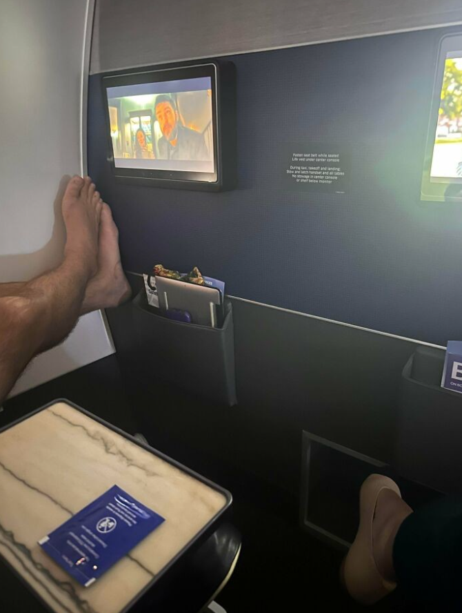 Cestujúci na palube lietadla sledujú film na obrazovke, zatiaľ čo si oddychujú so zdvihnutými nohami v priestore prvej triedy.