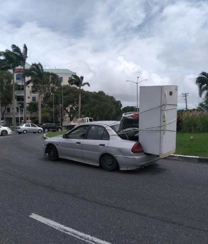 Auto s chladničkou na streche jazdí po ceste obklopenej palmami, neštandardná preprava.