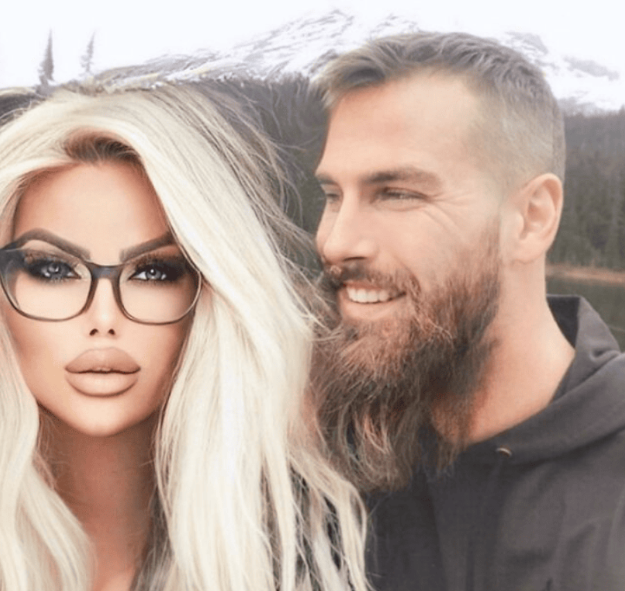 Pár na pozadí zasnežených hôr, muž s bradou a žena s blond vlasmi a okuliarmi v prírodnom prostredí.